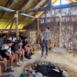 Alumnos sentados escuchan una explicación dentro de una cabaña de madera, junto a un hogar de piedras.