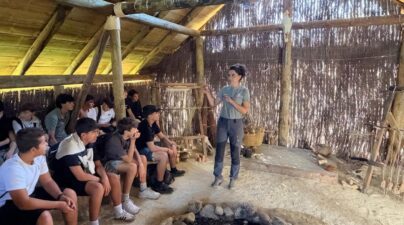 Alumnos sentados escuchan una explicación dentro de una cabaña de madera, junto a un hogar de piedras.