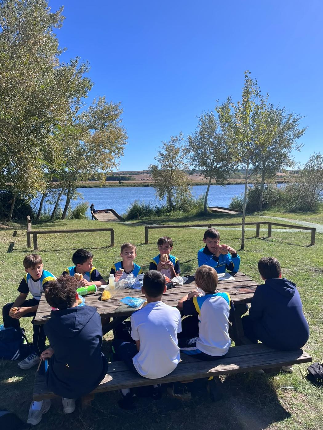 Varios alumnos comen sentados alrededor de una mesa de madera, junto al río y rodeados de árboles.