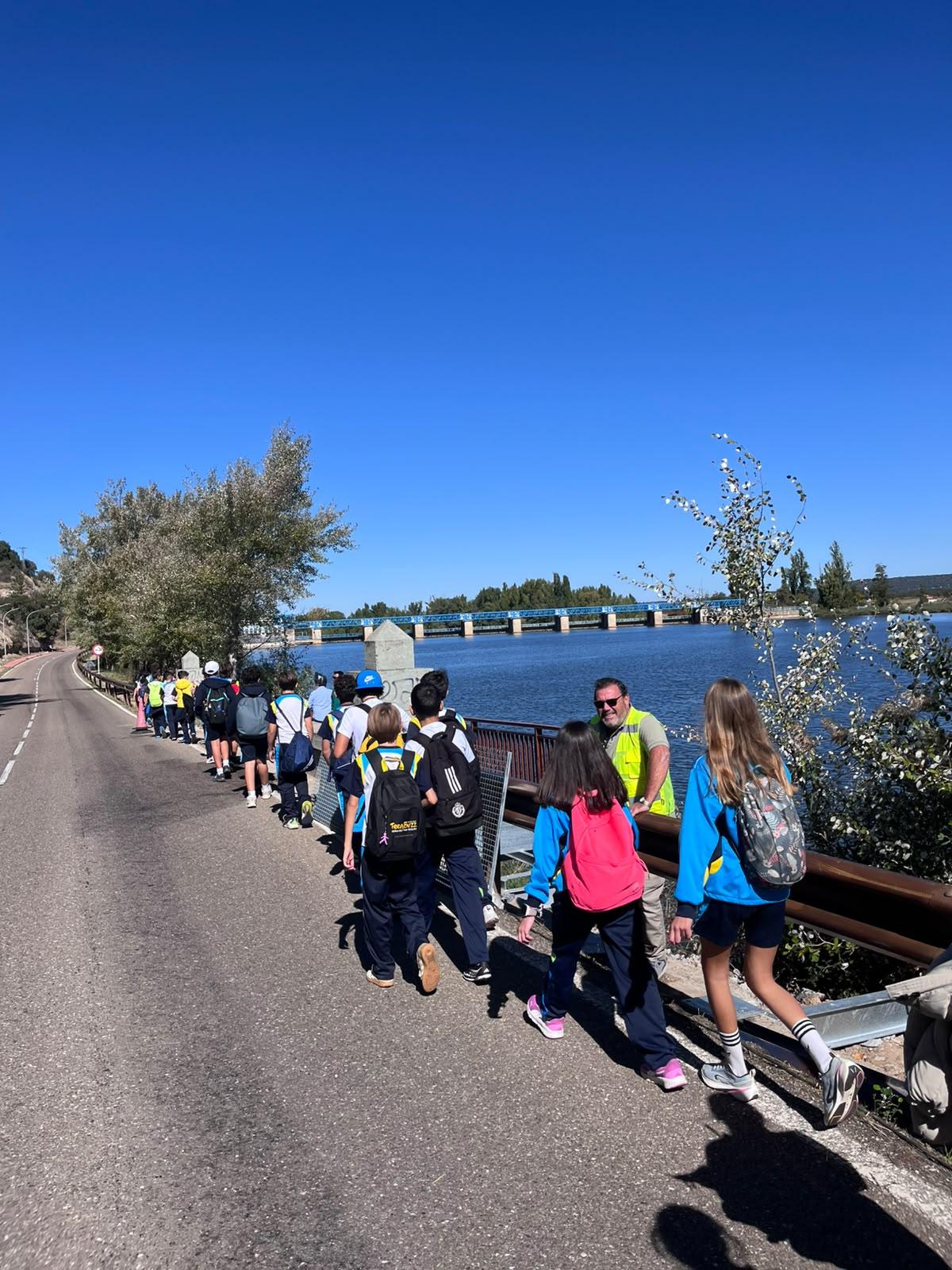 Los alumnos caminan en fila junto a la carretera, con el río y la presa de San José al fondo.