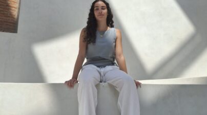 Joven sentada en unas gradas blancas, con un muro de ladrillo y grandes arcos al fondo.