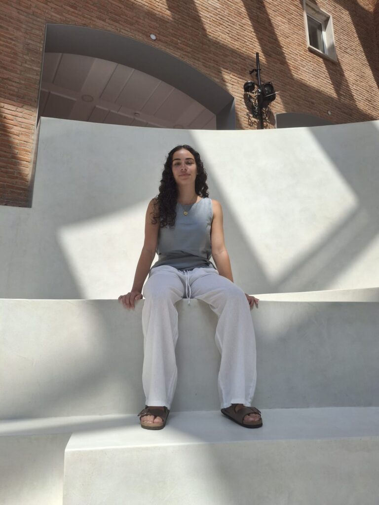 Joven sentada en unas gradas blancas, con un muro de ladrillo y grandes arcos al fondo.