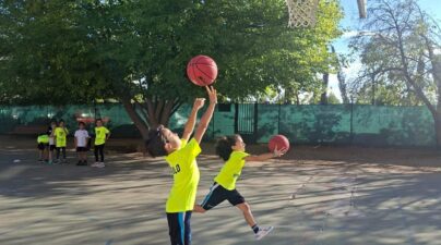 Dos niños con camisetas amarillas practican lanzamientos a canasta en el patio, con aros sobre el suelo.