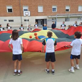 Alumnos sujetan en círculo un paracaídas de colores y hacen saltar varias pelotas sobre la tela en el patio.
