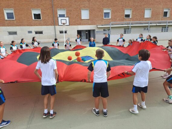 Alumnos sujetan en círculo un paracaídas de colores y hacen saltar varias pelotas sobre la tela en el patio.