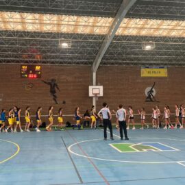 Jugadoras de ambos equipos se acercan para saludarse en la pista cubierta, con dos árbitros en primer plano.
