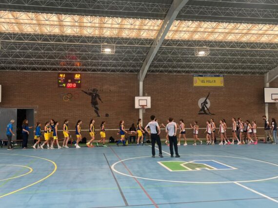 Jugadoras de ambos equipos se acercan para saludarse en la pista cubierta, con dos árbitros en primer plano.